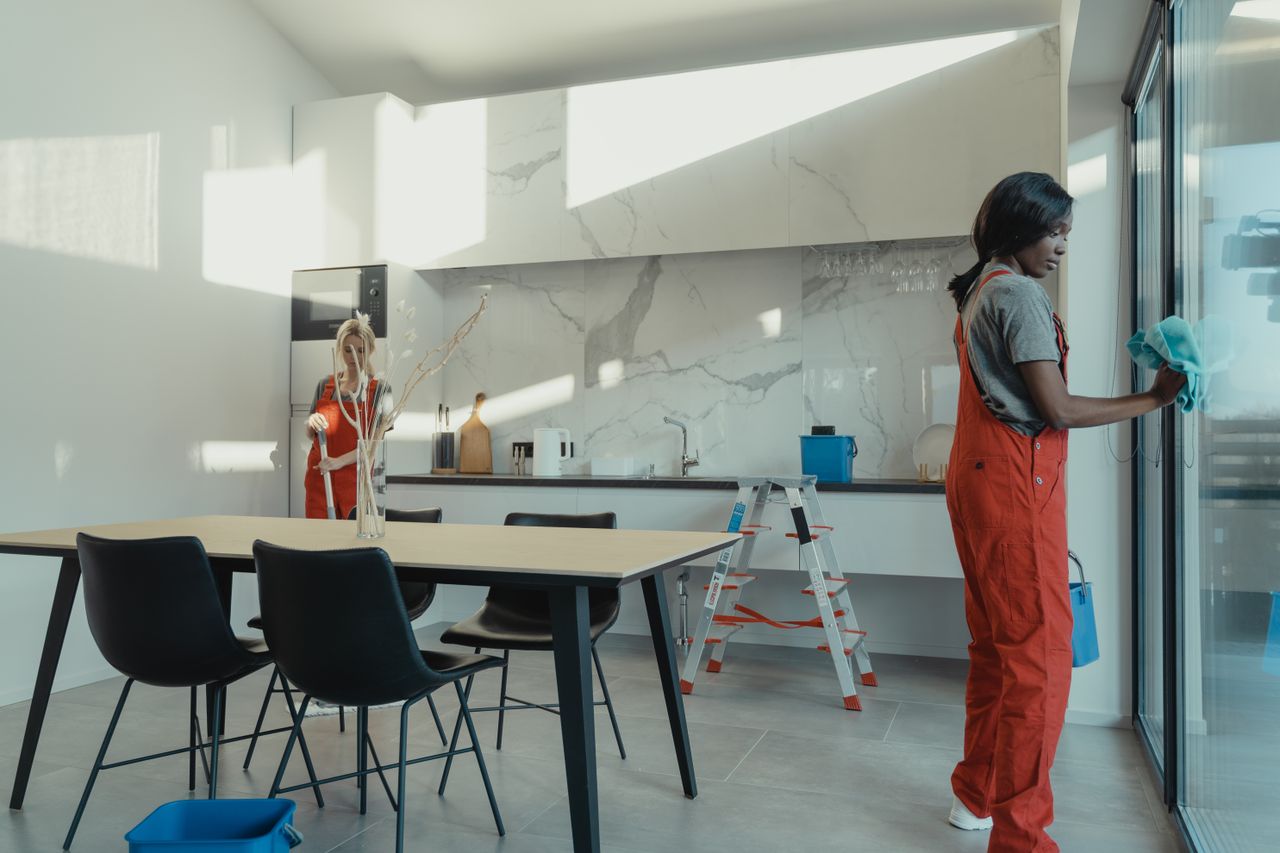 services-03 Two women in work clothes cleaning a kitchen with glass doors.